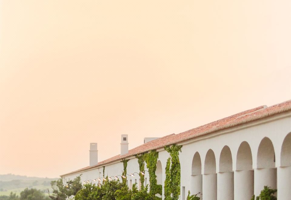 a long white building with columns and a red roof is surrounded by greenery , and the sky is orange at Sao Lourenco do Barrocal