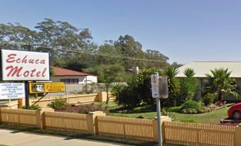 a sign for a motel is displayed in front of a fence and trees , with a house visible in the background at Echuca Motel
