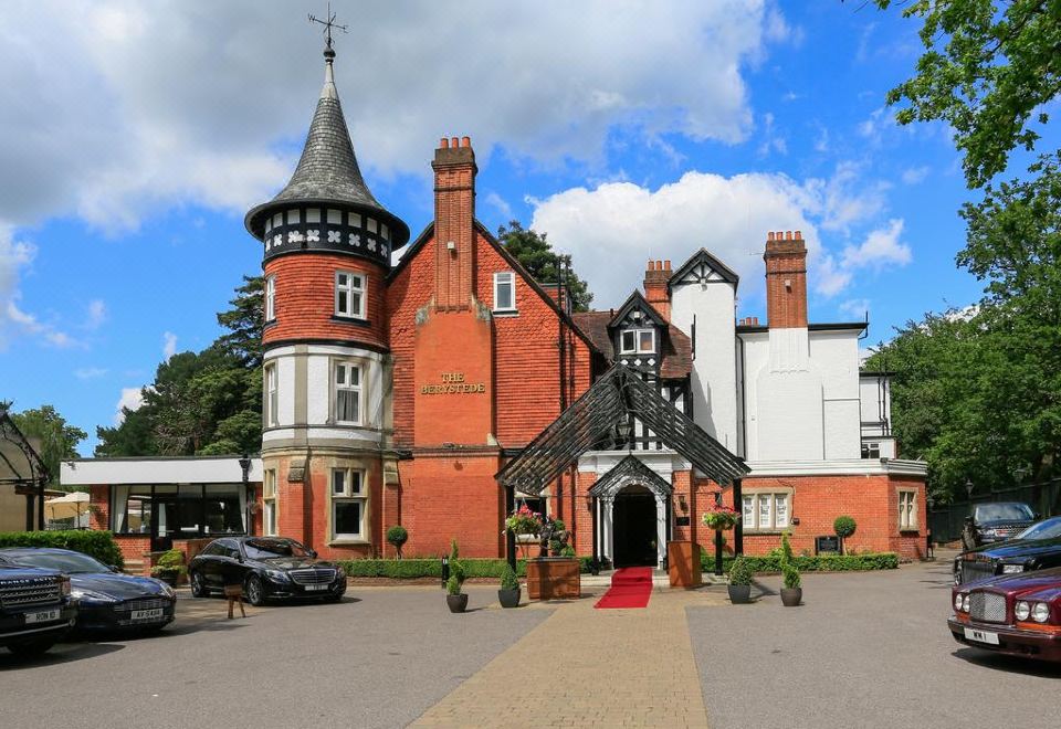 a large , red brick building with a white chimney and black roof , surrounded by trees and cars at Macdonald Berystede Hotel and Spa