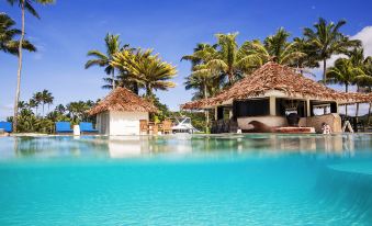 a large swimming pool with two thatched - roof huts in the background and palm trees surrounding it at The Pearl South Pacific Resort