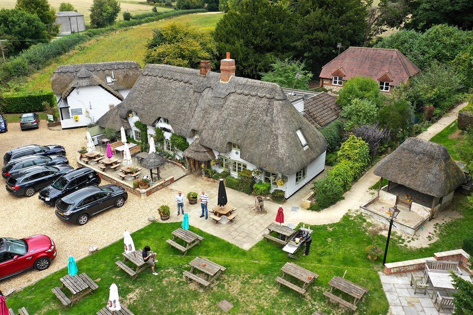 aerial view of a thatched - roof house surrounded by a grassy field , with people and vehicles in the background at The Hatchet Inn