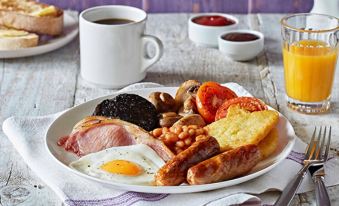 a plate of breakfast food , including ham , bacon , sausage , beans , eggs , and toast , sits on a table next to a cup at Premier Inn Lichfield North East (A38)