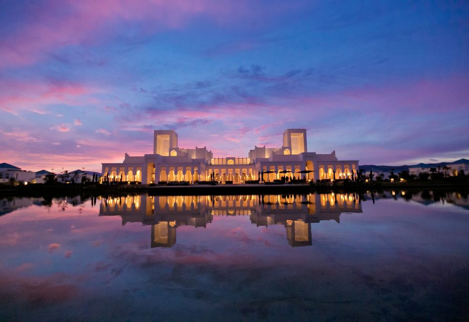 a large , white building with multiple floors and lit up windows is reflected in the water below at Banyan Tree Tamouda Bay