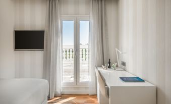 a modern hotel room with white curtains , a desk , and a window overlooking a balcony at NH Madrid Nacional