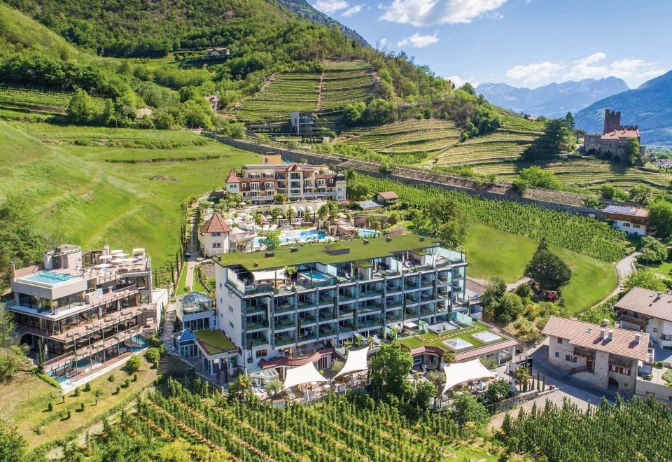 a large , multi - story building with a green roof is surrounded by vineyards and hills , all of which are lush with trees at Preidlhof Luxury Dolce Vita Resort