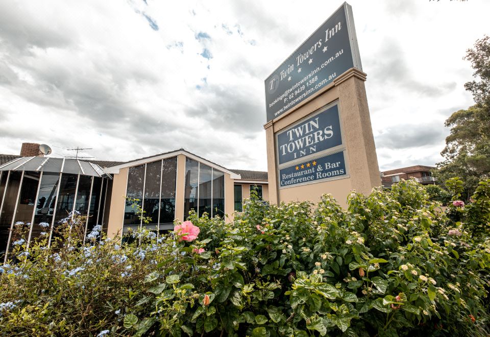 a building with a large sign on the front , surrounded by greenery and flowers , under a cloudy sky at Twin Towers Inn