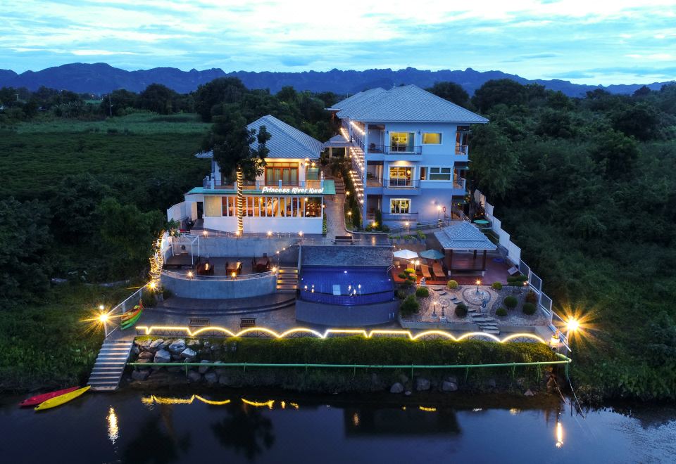 a large house situated next to a body of water , with a swimming pool in the background at Princess River Kwai Hotel