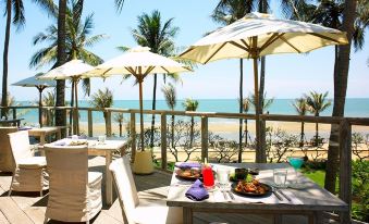 a beachfront restaurant with umbrellas , tables , and chairs set up on the sand , offering a panoramic view of the ocean at Anantara Hua Hin Resort