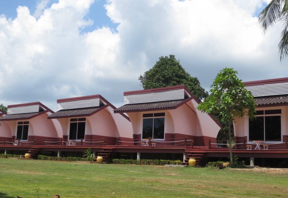 a row of small , pink houses situated on a grassy field , with trees and clouds in the background at Lomtalay Resort Trat