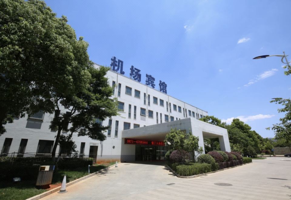 a large white building with a red sign on top , surrounded by trees and a parking lot at Airport Hotel