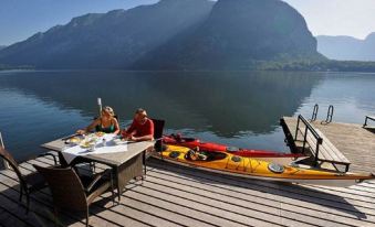 a couple is sitting at a dining table on a dock , enjoying a meal and each other 's company while a kayaker is floating nearby at Seehotel Grüner Baum
