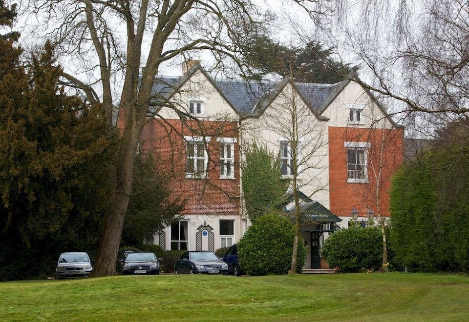 a brick house with two garages , situated in a grassy area with trees and a driveway at Coulsdon Manor Hotel and Golf Club