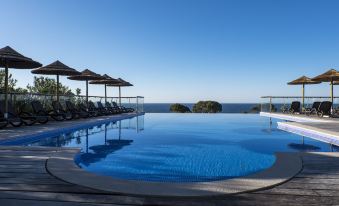 a large , oval - shaped swimming pool with clear blue water and umbrellas on the edge , overlooking a scenic ocean at Vila Alba Resort