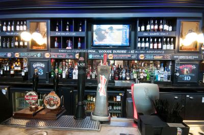 a bar with a tv screen displaying multiple bottles of alcohol and an electronic sign above it at Village Hotel Liverpool