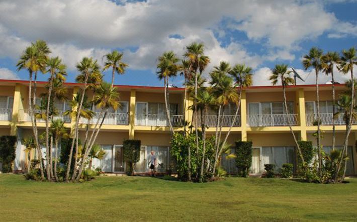 a large yellow building with palm trees in front of it , surrounded by green grass and trees at Colony
