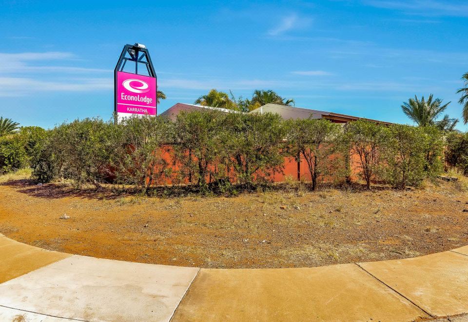 a red building with a pink sign on top , surrounded by trees and a dirt field at Econo Lodge Karratha
