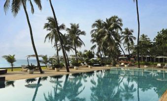 a large , clear blue swimming pool surrounded by palm trees and beach chairs , with the ocean visible in the background at Novotel Mumbai Juhu Beach