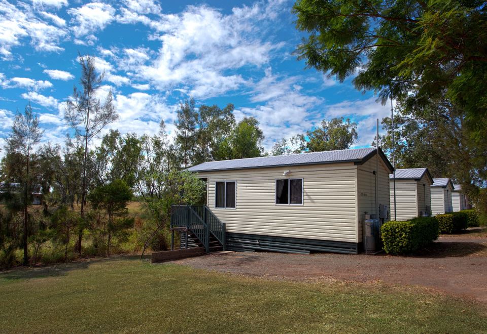 a small , white house with a metal roof and green roof is surrounded by trees and grass at Discovery Parks – Biloela