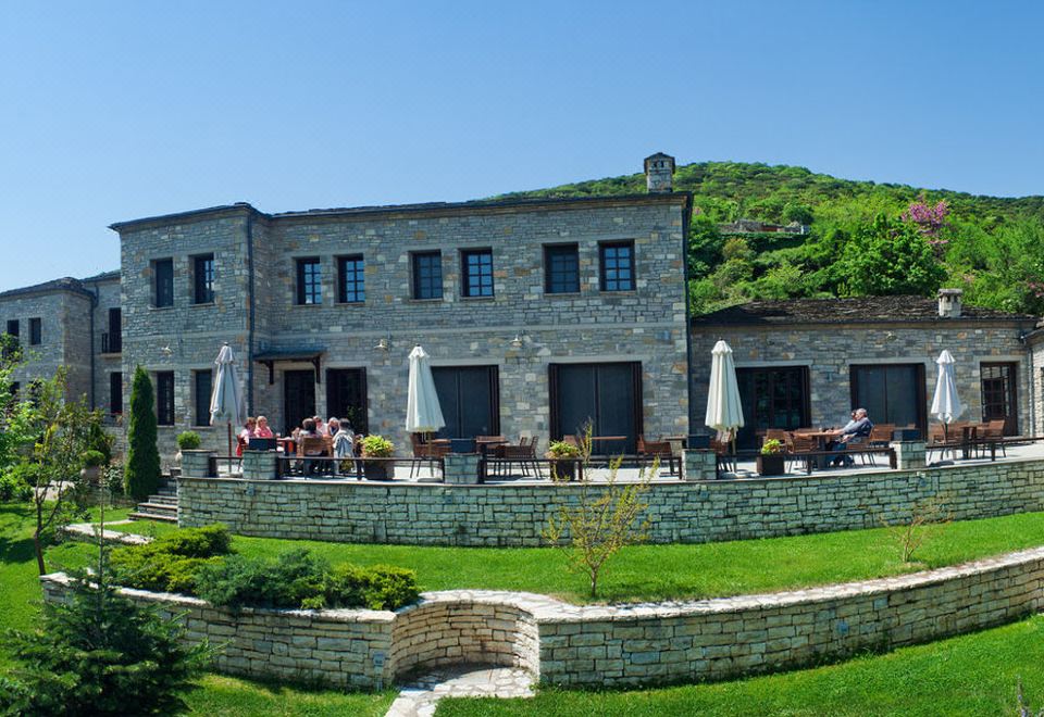 a stone building with a green lawn and people sitting at tables outside , surrounded by trees and mountains at Aristi Mountain Resort