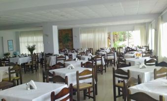 a large , empty dining room with wooden chairs and tables , white tablecloths , and windows covered by curtains at Paradise Beach Hotel