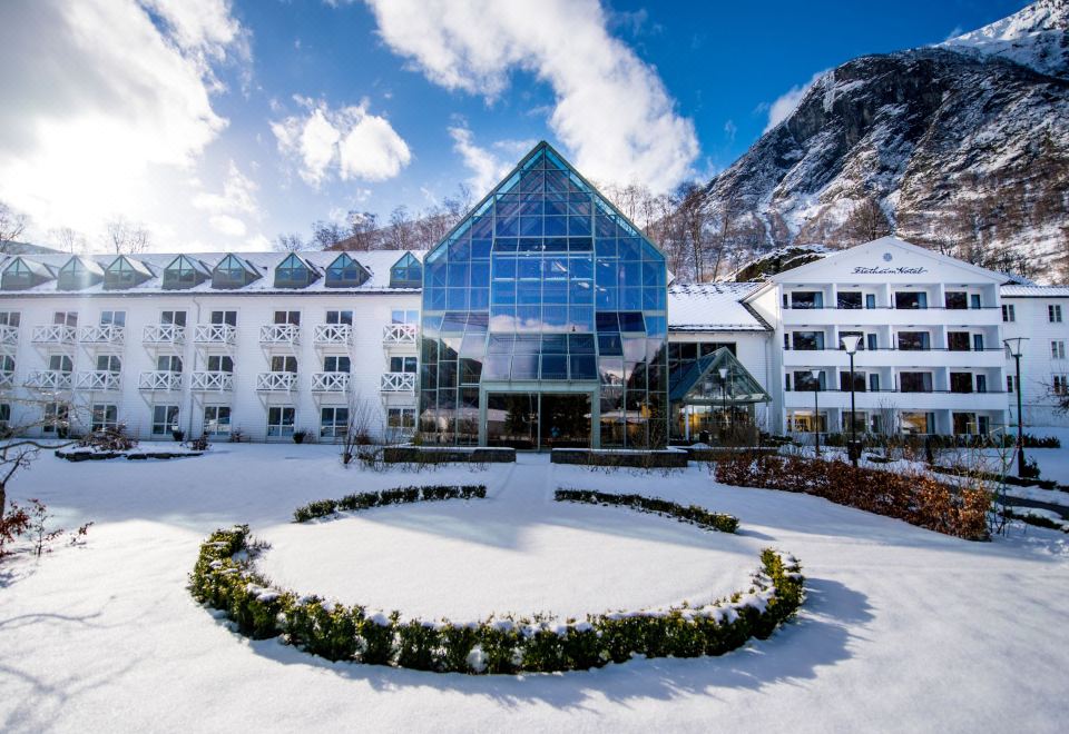 a large building with a blue glass exterior is surrounded by snow and has a circular flower bed in the front at Fretheim Hotel