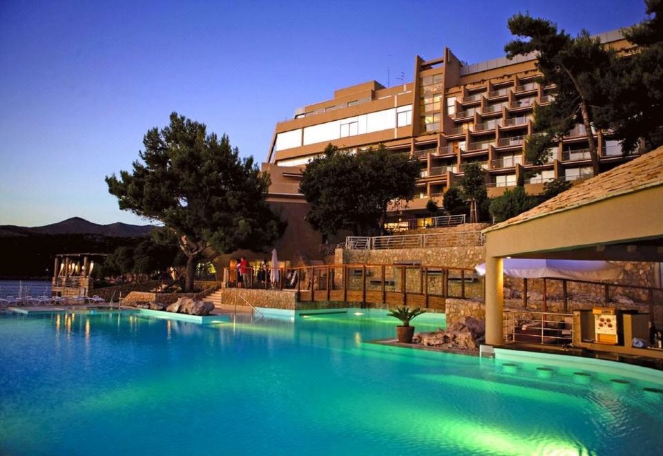 a large hotel with a pool and a bridge , surrounded by trees and buildings , under a clear blue sky at Hotel Dubrovnik Palace