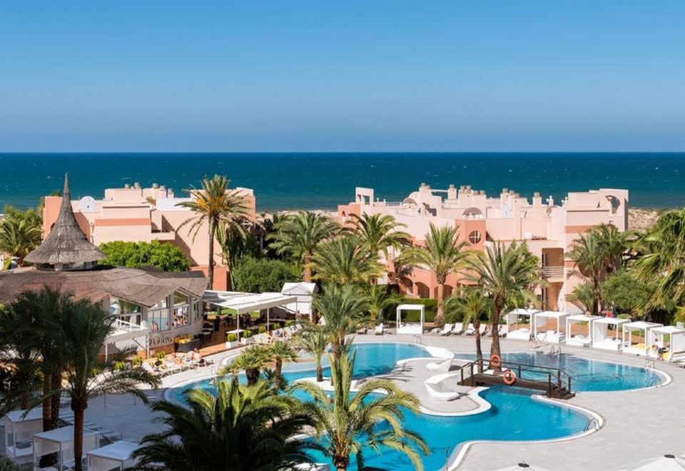 a resort with a pool surrounded by palm trees , chairs , and umbrellas , as well as a view of the ocean in the background at Oliva Nova Beach & Golf Hotel
