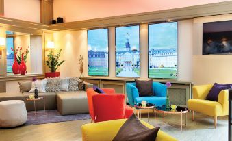 a brightly colored living room with multiple chairs and couches arranged in front of large windows at Leonardo Hotel Karlsruhe