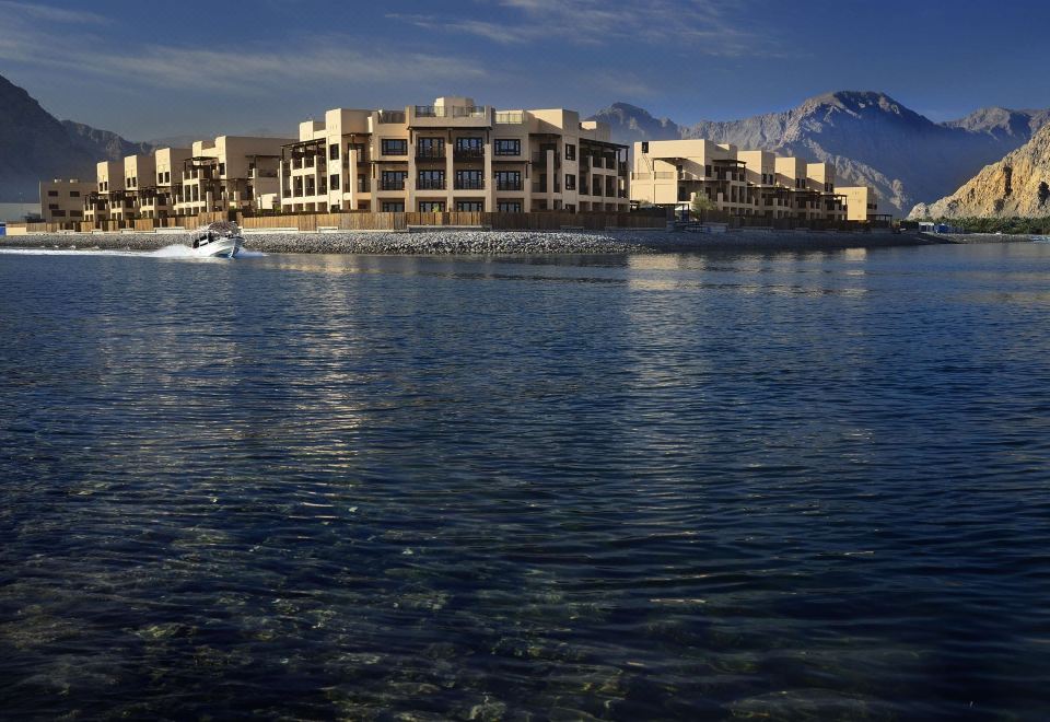 a large building is situated on the shore of a body of water , with mountains in the background at Atana Musandam Resort