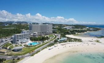 aerial view of a large white hotel on the beach , surrounded by palm trees and overlooking the ocean at ORION HOTEL MOTOBU RESORT&SPA