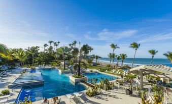a large outdoor pool surrounded by palm trees and lounge chairs , with a view of the ocean in the background at The St. Regis Bahia Beach Resort, Puerto Rico
