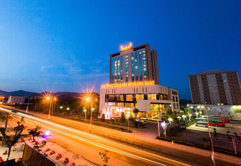 a tall hotel building with a yellow sign lit up at night , surrounded by trees and a road at Muong Thanh Grand Hoang Mai - Nghe An