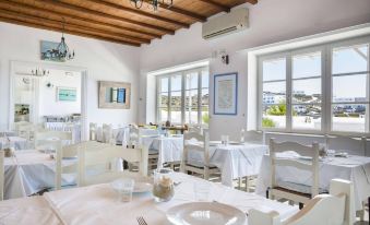 a white dining room with wooden ceiling and tables set for a meal , surrounded by windows that offer views of the ocean at Mykonos Beach Hotel