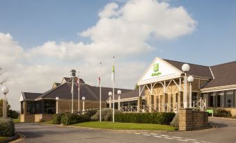 a large building with a green and white sign on the front , surrounded by trees at Holiday Inn Leeds - Brighouse