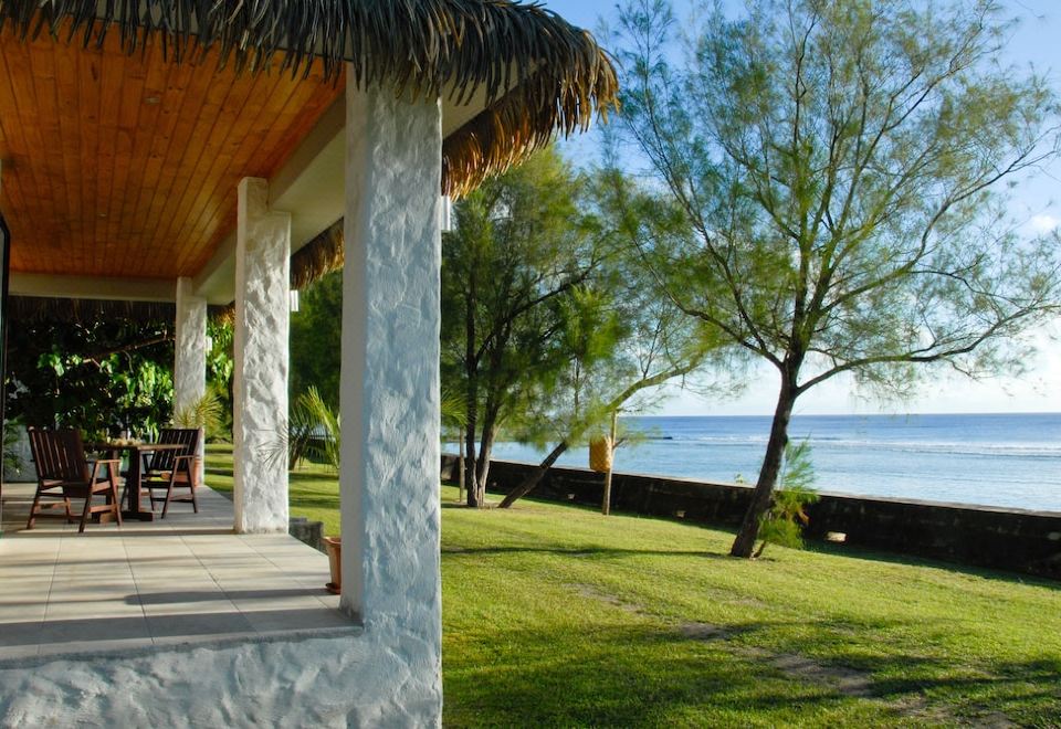 a porch with a thatched roof overlooks a grassy area and trees near the water at Mangaia Villas