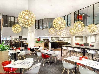 a modern restaurant with white tables and chairs , red accents , and large pendant lights hanging from the ceiling at Ibis Melbourne Hotel and Apartments