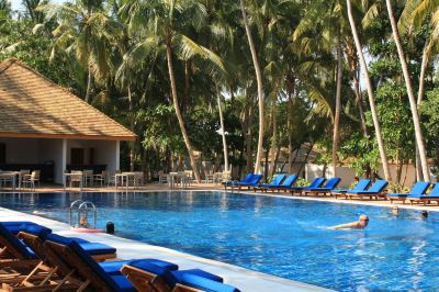 a large outdoor swimming pool surrounded by lounge chairs and palm trees , with people enjoying their time in the pool at Vilamendhoo Island Resort & Spa