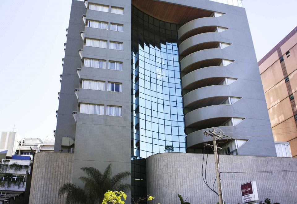 a modern building with a curved design and large windows is surrounded by palm trees at Mercure Belo Horizonte Vila da Serra