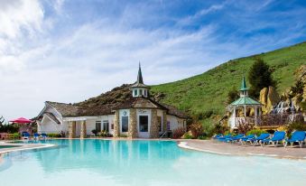 a large outdoor pool surrounded by green grass , with a house in the background and people relaxing by the pool at Madonna Inn