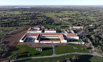 aerial view of a large white building surrounded by a grassy field , with a golf course in the background at São Lourenço do Barrocal