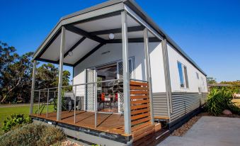 a small , modern house with a white exterior and wooden deck , surrounded by trees and greenery at Nrma Halls Gap Holiday Park