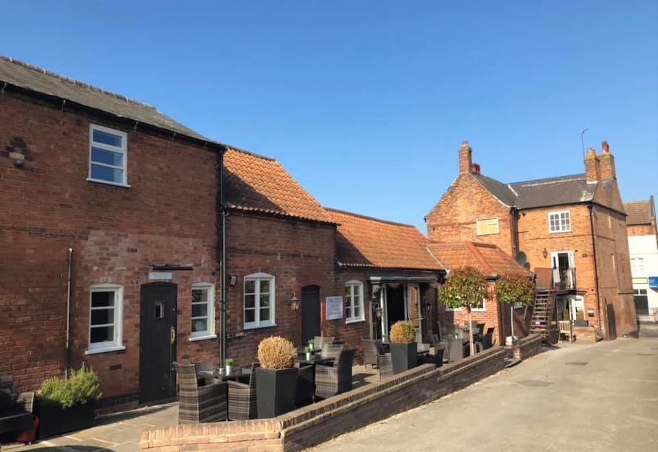 a brick building with a red roof and windows is lined with potted plants on the sidewalk at The Wheatsheaf Pub, Kitchen & Rooms