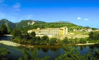 a large building with a long row of windows is situated on the bank of a river , surrounded by greenery and mountains at Parador de Cangas de Onis