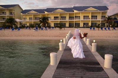 a bride in a white wedding dress is walking on a dock near the water , with a hotel in the background at Wyndham Reef Resort Grand Cayman