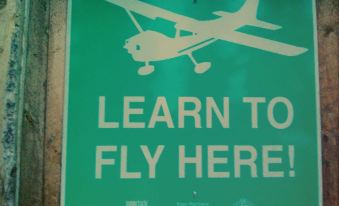 "a green sign with white text that reads "" learn to fly here !"" and a small plane flying above it" at Pine Point Resort