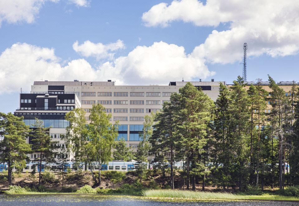 a large building with a blue and white facade is surrounded by trees and a body of water at Quality Hotel Winn Haninge