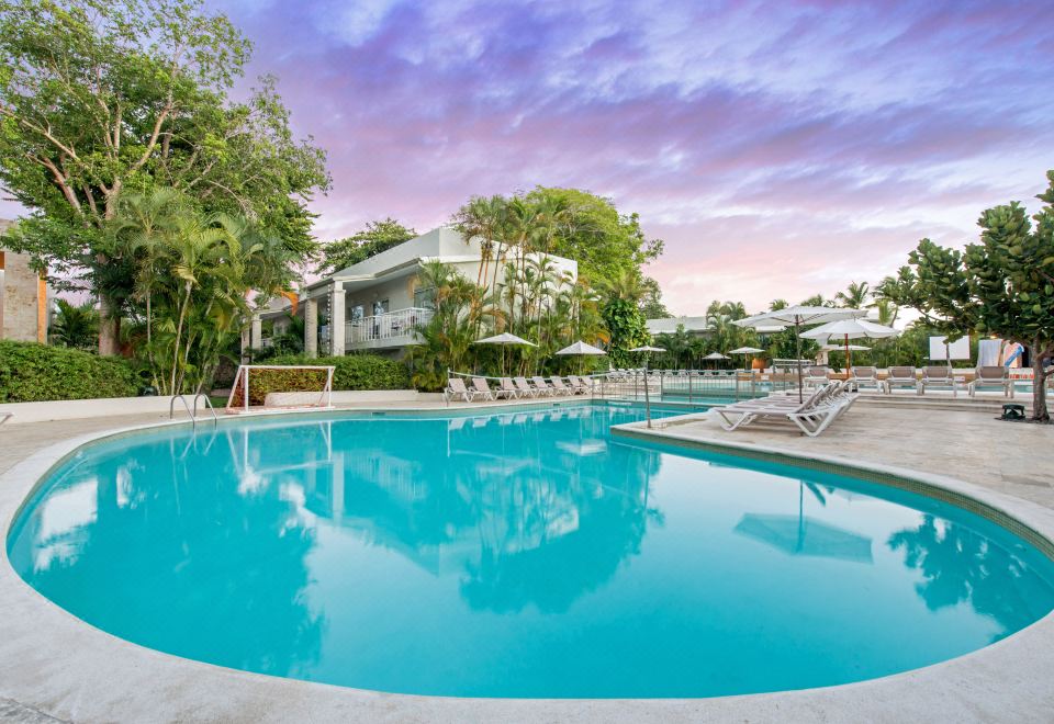 a large outdoor swimming pool surrounded by lounge chairs and umbrellas , with a house in the background at Emotions by Hodelpa - Juan Dolio