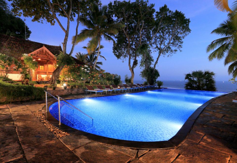 a large blue swimming pool surrounded by palm trees and a house , with the ocean visible in the background at Somatheeram Ayurveda Village