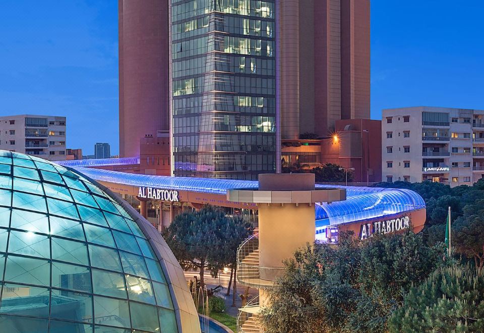 a tall building with a glass dome and blue lights , surrounded by trees and a circular structure at Hilton Beirut Metropolitan Palace