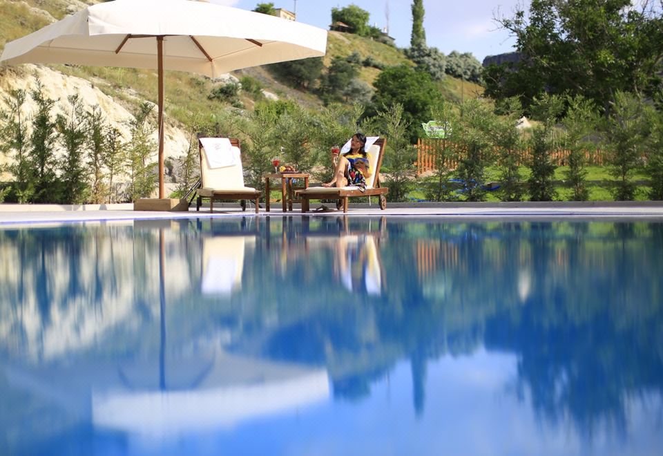 a woman is relaxing by the edge of a large blue pool , surrounded by chairs and an umbrella at Kapadokya Hill Hotel & Spa (12+)